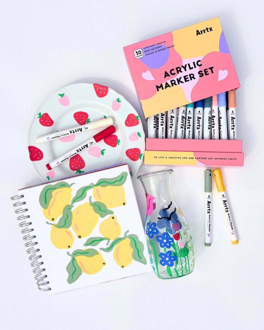 Arrtx acrylmarker set met 30 kleuren, naast eindresultaten: beschilderd bordje met aardbeien, schetsboek met citroenen en een glazen karaf met bloemen